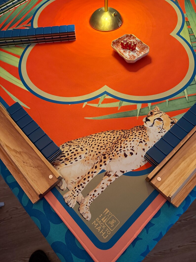 May include: Mahjong set with a vibrant orange and blue floral design, featuring a cheetah illustration. The set includes blue tiles, wooden racks, and a small dish with red dice. The game is set on a patterned mat.
