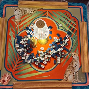 May include: A colorful Mahjong game set on a table with a vibrant orange and teal design. The table features illustrations of cheetahs and tropical foliage. The game pieces are blue and white, and a decorative lamp sits in the center.