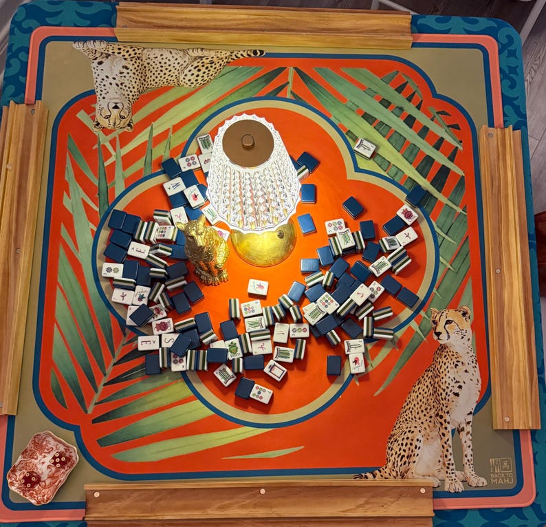 May include: A colorful Mahjong game set on a table with a vibrant orange and teal design. The table features illustrations of cheetahs and tropical foliage. The game pieces are blue and white, and a decorative lamp sits in the center.
