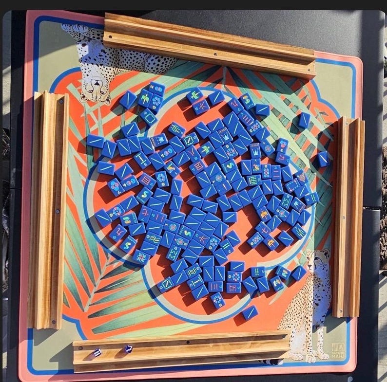 May include: A Mahjong set with blue tiles scattered on a colorful game board. The board features a tropical design with orange, green, and pink hues, and wooden tile holders. The board has a cheetah print border.
