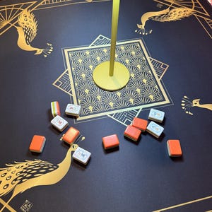 Puede incluir: Un tapete de mahjong negro con ilustraciones de pavos reales dorados y patrones geom&eacute;tricos, con fichas de mahjong dispersas que muestran dorsos naranjas y caras blancas con caracteres azules. Una base de l&aacute;mpara dorada se encuentra en el centro.