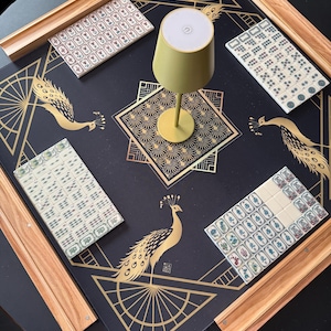Puede incluir: Una alfombrilla de mahjong cuadrada de color negro con patrones geom&eacute;tricos y de pavos reales dorados, cuatro soportes de madera y juegos de fichas blancas. Una peque&ntilde;a l&aacute;mpara de color oliva con la parte superior blanca est&aacute; en el centro.