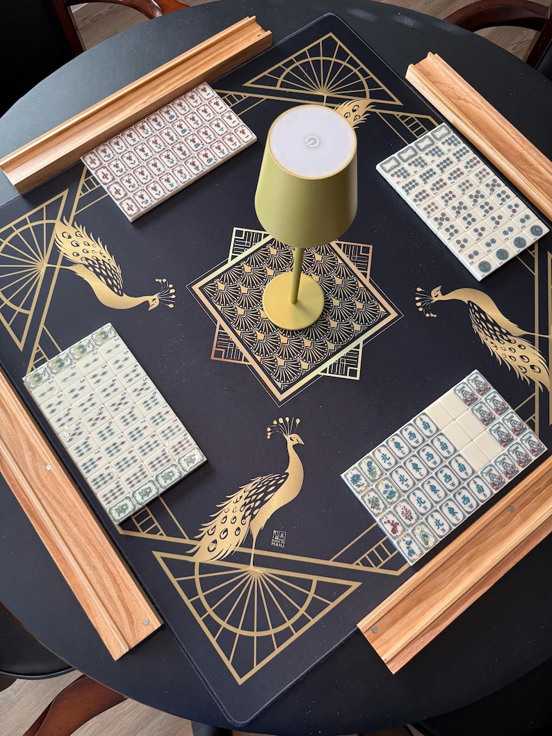 Puede incluir: Una alfombrilla de mahjong cuadrada de color negro con patrones geom&eacute;tricos y de pavos reales dorados, cuatro soportes de madera y juegos de fichas blancas. Una peque&ntilde;a l&aacute;mpara de color oliva con la parte superior blanca est&aacute; en el centro.