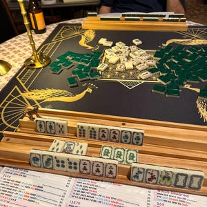 Puede incluir: Un juego de Mahjong con fichas color marfil y reverso verde, dispuestas sobre un tapete negro con ilustraciones de pavos reales dorados. Incluye soportes de madera, una tarjeta de puntuaci&oacute;n con texto impreso y una l&aacute;mpara dorada.