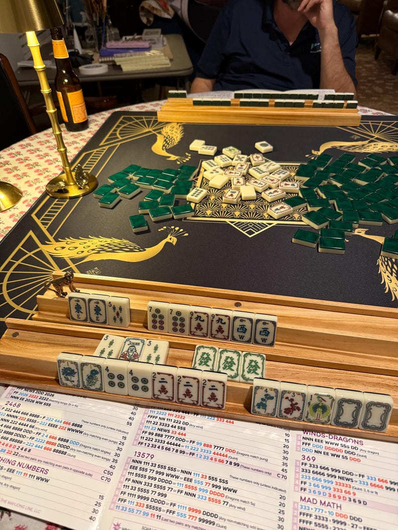 Puede incluir: Un juego de Mahjong con fichas color marfil y reverso verde, dispuestas sobre un tapete negro con ilustraciones de pavos reales dorados. Incluye soportes de madera, una tarjeta de puntuaci&oacute;n con texto impreso y una l&aacute;mpara dorada.