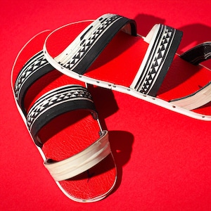 Puede incluir: Un par de sandalias rojas con correas estampadas en blanco y negro. Las sandalias tienen una suela plana y un diseño sencillo, perfectas para uso casual. El fondo es rojo liso.