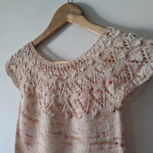May include: A cream-colored, hand-knitted top with a scoop neck and short sleeves. The top features a delicate openwork design around the neckline and sleeves, with subtle pink and yellow flecks throughout the fabric. The garment is displayed on a wooden hanger.