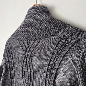 Puede incluir: Un suéter gris tejido con un cuello alto y detalles intrincados de punto de cable. El suéter está colgado en una percha de madera contra una pared blanca. El diseño presenta patrones verticales y diagonales.