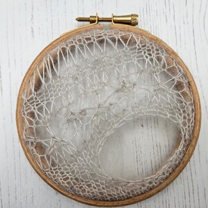 May include: A handmade, circular embroidery hoop with a natural wood finish. The hoop holds a delicate, web-like design crafted from off-white thread, creating a textured, open pattern. The top features a brass screw for tension adjustment.