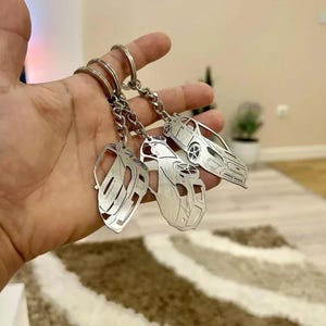 May include: Three silver car-shaped keyrings with detailed cut-out designs. Each keyring showcases a different car silhouette, connected to a chain and a ring. The keyrings are held in a hand.