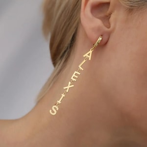 May include: Gold-coloured ALEXIS name earrings. The earrings feature the name ALEXIS spelled out in individual letters that dangle from a hoop. The letters are connected by small links. The earrings are worn on the ear.