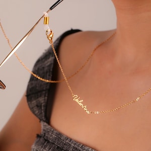 May include: A gold-coloured eyeglass chain with the name "Valentina" in cursive script. The chain is attached to the glasses with a small gold loop. The chain is worn around the neck.