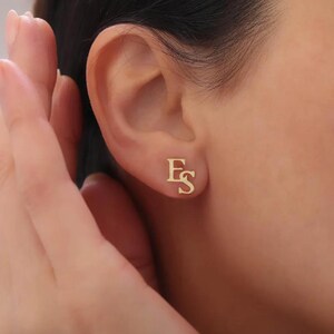 May include: Gold-coloured stud earrings featuring the letters "E" and "S" in a decorative font. The earrings are small and positioned on the earlobe. The background is blurred, highlighting the jewellery.