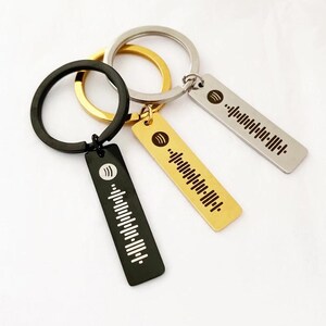 May include: Three metal keyrings with Spotify code designs. One is black, one is gold, and one is silver. Each keyring has a rectangular tag with a unique audio waveform pattern and the Spotify logo. The keyrings are gold, silver, and black.