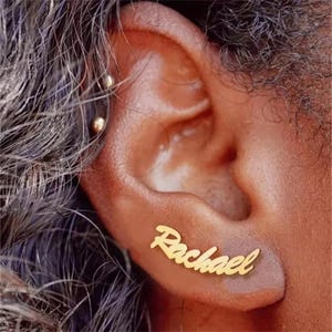 May include: Gold-coloured "Rachael" name earrings in a cursive font, worn on the ear. The image is a close-up of the ear with the earrings in place. A small gold stud earring is also visible in the image.