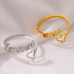 May include: Two open-style rings, one silver and one gold, each featuring a name and a heart-shaped accent. The silver ring spells "Emma", while the gold ring spells "Emma". The rings are displayed on a soft, light-coloured surface.