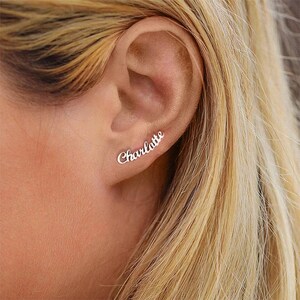 May include: Silver ear climber earring spelling the name "Charlotte" in a cursive script. The earring is worn on the ear, with the ear and blonde hair visible in the background.