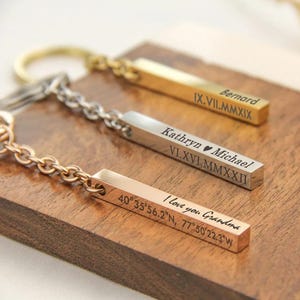 May include: Three personalised bar keyrings in gold, silver, and rose gold. Each bar is engraved with text, including names, dates, and a message. The keyrings are displayed on a wooden surface.