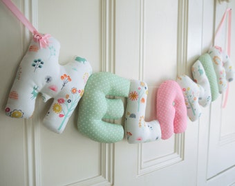 May include: A nursery banner with the name "MELANIE" spelled out in colorful fabric letters. The letters are sewn together with pink ribbon and feature a variety of floral and animal patterns.