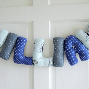 May include: A blue and gray fabric letter banner spelling the name "WILLIE". Each letter is made of soft fabric with a different pattern, including blue polka dots, gray polka dots, and a light blue pattern with sailboats and beach houses.