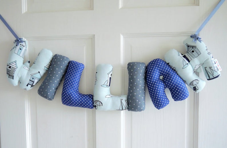 May include: A blue and gray fabric letter banner spelling the name "WILLIE". Each letter is made of soft fabric with a different pattern, including blue polka dots, gray polka dots, and a light blue pattern with sailboats and beach houses.