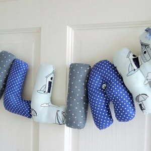 May include: A blue and white fabric banner with the name "WILLIAN" spelled out in individual letters. Each letter is decorated with a nautical theme, featuring small white sailboats, lighthouses, and beach houses.