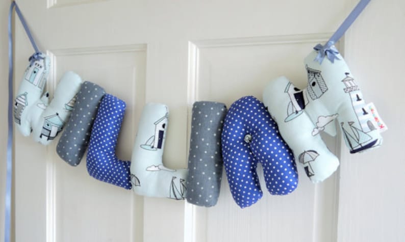 May include: A blue and white fabric banner with the name "WILLIAN" spelled out in individual letters. Each letter is decorated with a nautical theme, featuring small white sailboats, lighthouses, and beach houses.