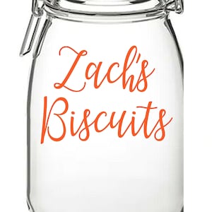 Może przedstawiać: Szklany słoik z przezroczystego szkła z zawiasem i etykietą z napisem "Zach's Biscuits" w pomarańczowych literach.