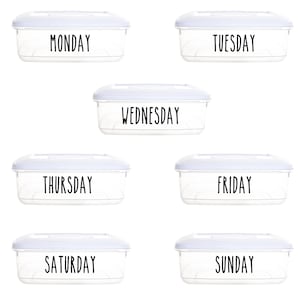 Op de afbeelding: Zes doorzichtige plastic containers met witte deksels, gelabeld met de dagen van de week: maandag, dinsdag, woensdag, donderdag, vrijdag, zaterdag en zondag.