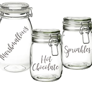 Op de afbeelding: Drie glazen potten met luchtdichte deksels en etiketten. De etiketten zijn in cursief geschreven en zeggen "Marshmallows", "Hete Chocolade" en "Sprinkels".