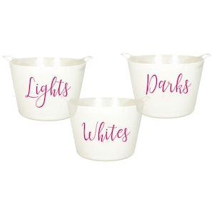 Può includere: Tre cesti per la biancheria in plastica bianca con scritte rosa. I cesti sono etichettati "Lights", "Darks" e "Whites".