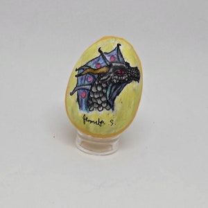May include: A hand-painted rock with a black dragon head with red eyes and a blue and purple wing design on a yellow background. The rock is on a clear stand. The artist's signature "Jennifer S." is written on the rock.