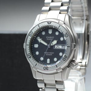 May include: A silver-toned stainless steel Casio Quartz wristwatch featuring a dark blue circular dial with luminous hour markers, a day-date display, and a rotating bezel. The watch includes a metal link bracelet and is marked Water Resist 20 Bar.
