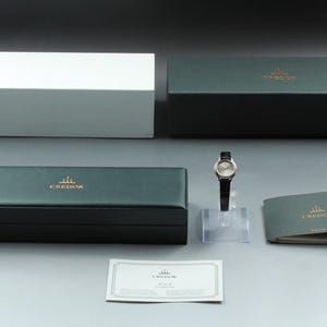 May include: A Credor wristwatch with a round silver dial and a black leather strap, displayed with a dark green presentation box, a white outer box, a matching green instruction booklet, and a white rectangular warranty card with the Credor logo.