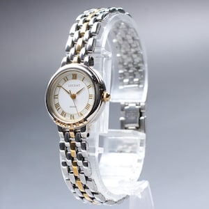 Puede incluir: Un reloj de pulsera analógico redondo con esfera blanca, números romanos dorados y el texto LUCENT impreso en la esfera. El reloj cuenta con una correa de eslabones metálicos bicolor que combina segmentos de metal pulido plateado y dorado.