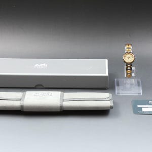May include: A silver and gold-tone wristwatch with a round cream-colored face, featuring a metallic link bracelet. Included are a gray rectangular box, a gray fabric watch roll, a small information card, and a dark gray warranty card with Hermes text.