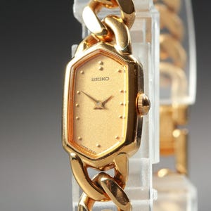 May include: A gold-tone Seiko wristwatch featuring a hexagonal case and a textured gold dial with simple dot hour markers. The watch has a thick, polished gold-tone chain link bracelet and a small crown on the right side of the case.