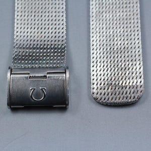 May include: A silver-toned metal watch strap featuring a fine mesh weave pattern. The strap includes a rectangular clasp mechanism engraved with the Greek letter Omega symbol, which serves as a functional closure for the watch band accessory.