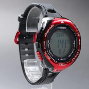 May include: A Seiko solar digital watch with a black rubber strap and a metallic red bezel. The round display shows the time, day, and date. The bezel features white text labels including Mode/Select, Light, Reset, Start/Stop, Solar, and Sensor.