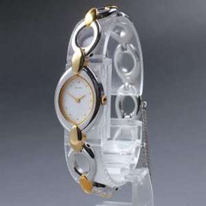 May include: A Seiko wristwatch featuring a white oval dial with gold-tone hands and markers. The watch has a two-tone bracelet composed of alternating silver-tone circular links and polished gold-tone oval accents, secured with a clasp.