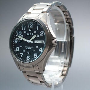 May include: Silver-toned ALBA titanium watch with a black face. The watch features white numerals, hands, and the date. The watch band is made of matching silver metal links. The watch is displayed on a clear stand.