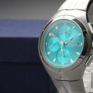 May include: A silver-toned stainless steel chronograph wristwatch featuring a vibrant turquoise dial with three sub-dials, silver hour markers, a date window at the three o'clock position, and the text WIRED printed on the watch face.