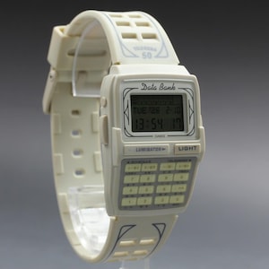 May include: A cream-colored Casio Data Bank watch with a calculator keypad and digital display. The watch band and face are off-white, with the time displayed as 13:54. The watch has the text "Data Bank" and "Luminator" printed on the face.