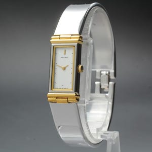 May include: A rectangular Seiko wristwatch featuring a white dial with gold-tone hands and markers. The watch case is silver-tone with gold-tone accents at the top and bottom. It has a polished silver-tone metal bangle-style bracelet band.
