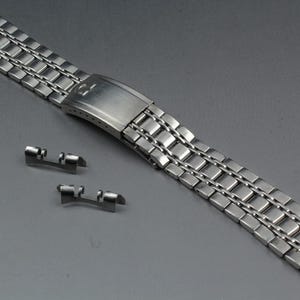 Peut inclure: Un bracelet de montre en acier inoxydable avec un design à maillons rectangulaires. Le bracelet est présenté avec deux barrettes à ressort. Le fermoir est visible et a une finition brossée. Le bracelet mesure environ 19 cm de long.