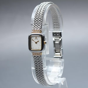 May include: A silver-tone Credor wristwatch featuring a square-shaped case with rounded corners, a white dial, and a textured woven metal link bracelet. The watch includes gold-tone accents at the lugs, which are set with small clear stones.