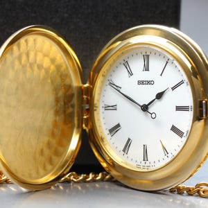 May include: A gold-tone pocket watch with an open hinged cover featuring a textured interior. The white dial displays black Roman numeral hour markers, black hands, and the Seiko brand name. A gold-tone chain is attached to the side of the watch.