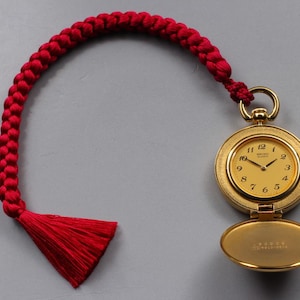 May include: A gold-tone pocket watch with a round face, black numerals, and thin hands. The watch features a hinged cover and is attached to a thick, braided red cord that terminates in a matching red tassel, creating a decorative accessory.