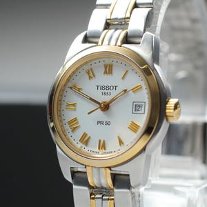 May include: A Tissot 1853 PR 50 wristwatch featuring a white dial with gold-tone Roman numeral hour markers and hands. The watch has a two-tone stainless steel and gold-tone metal link bracelet, a round gold-tone bezel, and a date display window.