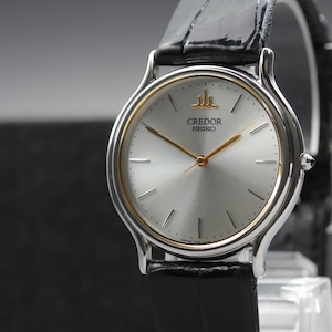 May include: A Credor Seiko wristwatch featuring a round silver dial with gold-tone hands and hour markers. The watch has a polished silver-tone case with a thin gold-tone inner bezel and a black leather strap with a textured, glossy finish.